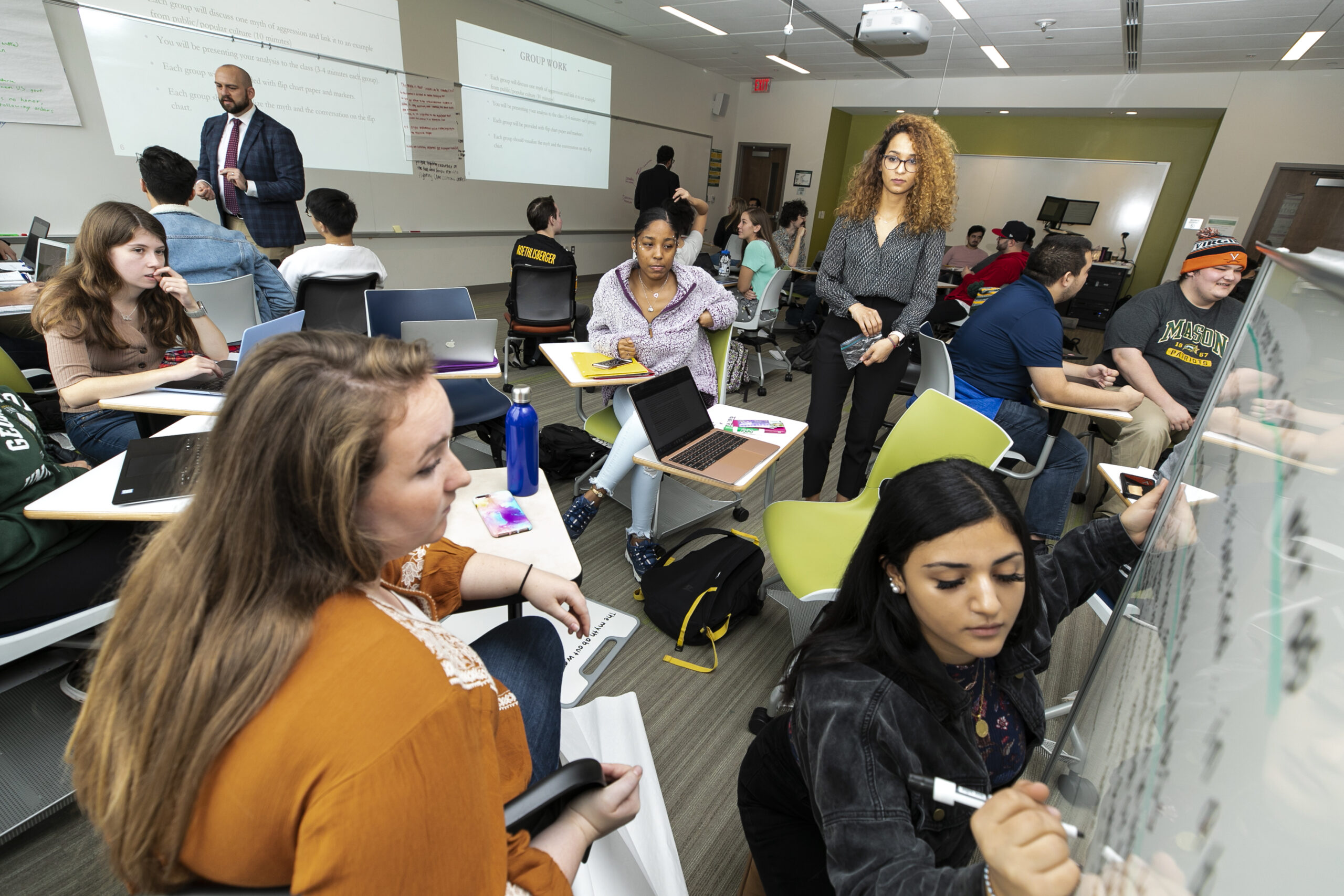 A student writes on a whiteboard while other students and instructor look on.